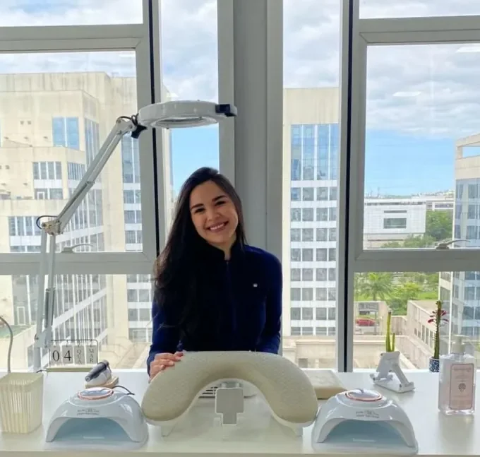 Fernanda Mezine sorrindo na mesa de atendimento do estúdio, com as 2 cabines de LED ao lado.